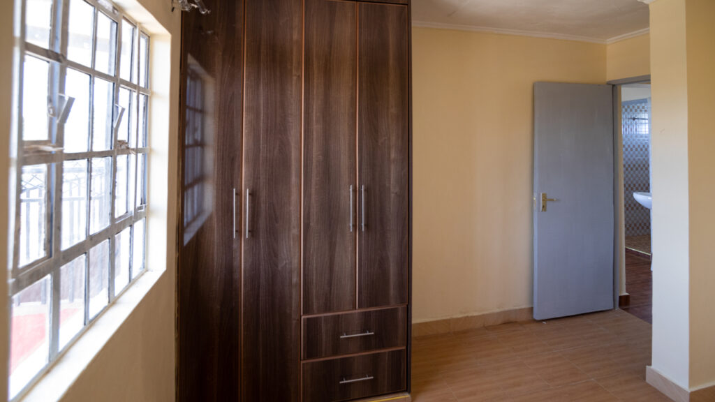 A well-lit room with a wooden wardrobe, a window allowing natural light, and a blue door.